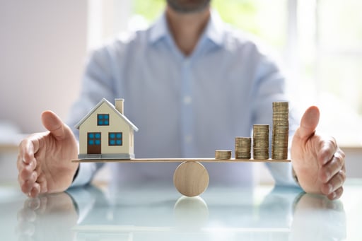 Small House Model and Stack of Gold Coins Balancing On Wooden Seesaw
