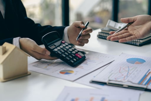 Real estate broker talks to client while holding calculator over table with projections and home model