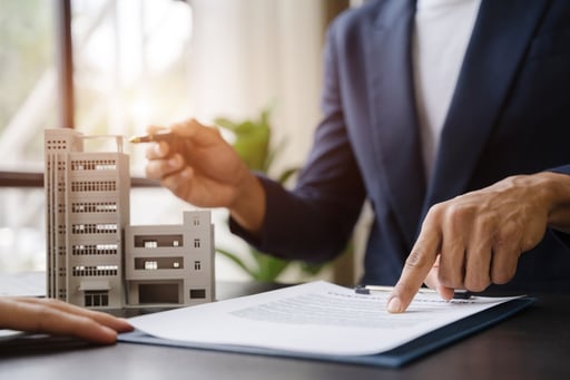 Real estate broker pointing to contract on table next to model of apartment building while talking to client