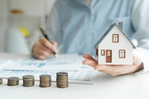 Man holding model of home with growing stacks of coins and loan papers on a table