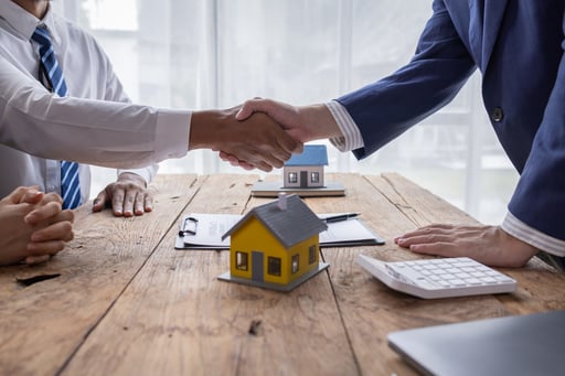 Real estate broker shaking hands with client over table with agreement, calculator, and model of homes
