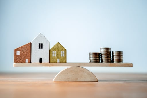 Scale with small wood homes on one side and coins money on the other side