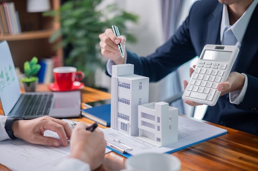 Real estate broker guiding client through property investment at table with models of homes and calculator in hand