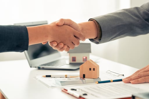 Close-up of a handshake between a real estate agent and a buyer over table with agreement and models of homes