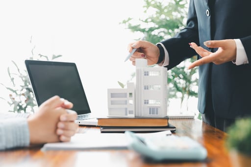 Real estate broker meets with client at desk with laptop calculator and model of apartment building