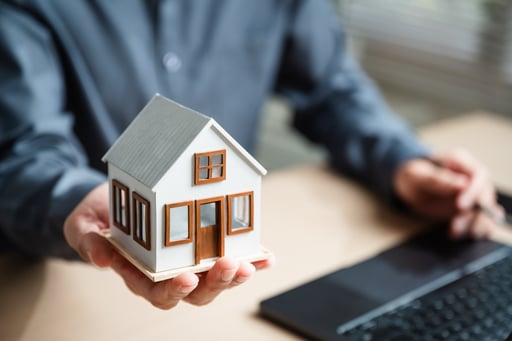 Broker holding a small model house in his hand while sitting at a desk with a laptop.