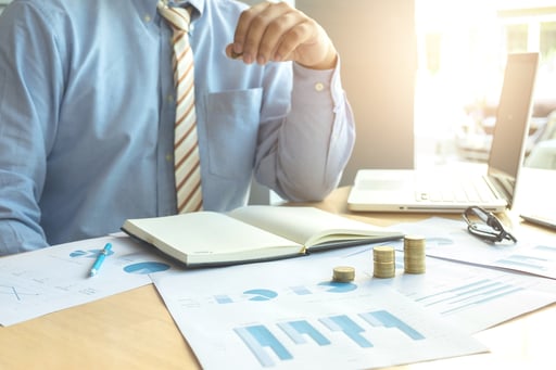 businessman pointing to ledger and graphs on table with laptop and stacks of growing coins